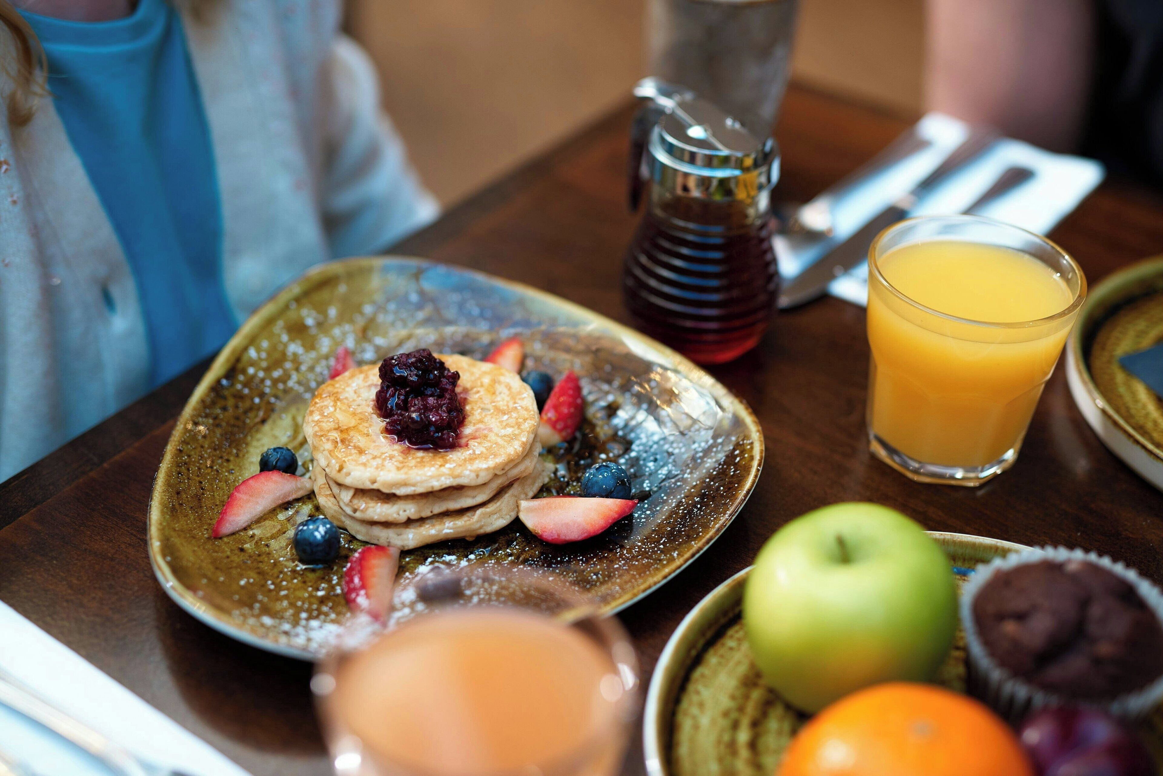Un desayuno con tortitas, fruta, un vaso de zumo de naranja y una jarra de sirope sobre una mesa.