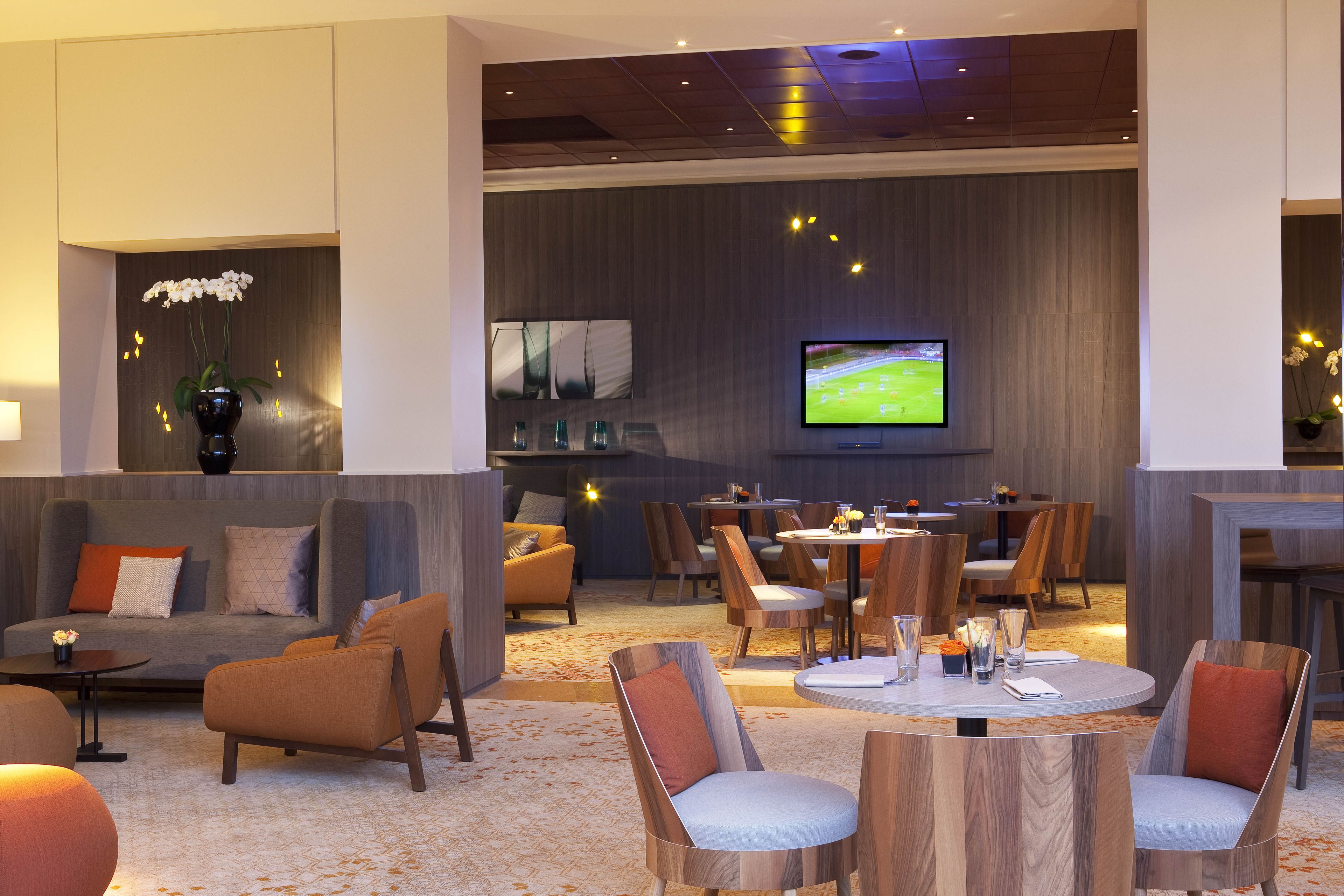 A modern hotel lobby with a seating area, a TV screen, and a bar.