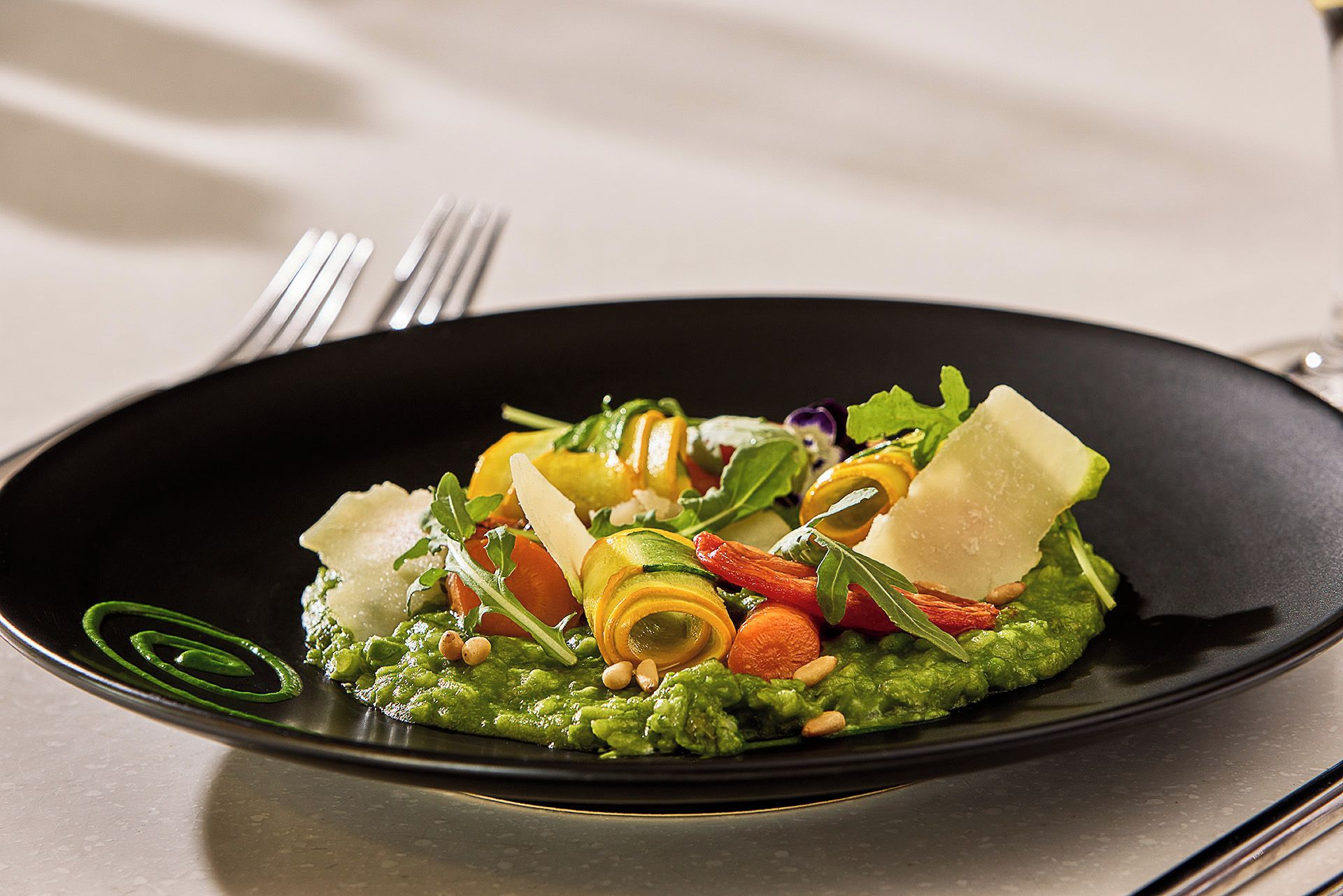 A black plate with a green vegetable puree, sliced vegetables, nuts, and cheese.