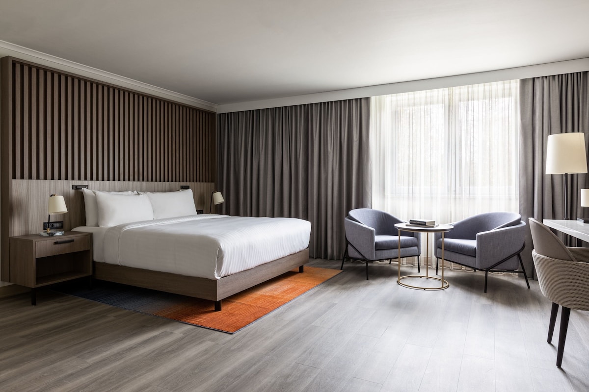 Et moderne hotelværelse med en stor seng, to lænestole, et lille bord og et skrivebord med en lampe.