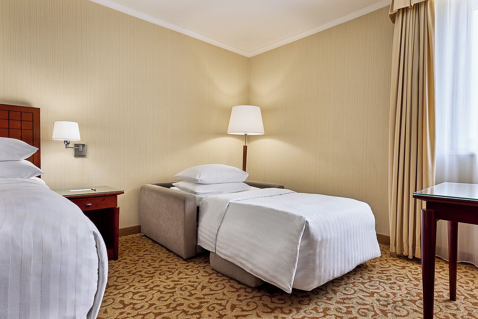 A hotel room with two beds, a desk, a lamp, and a window with curtains.
