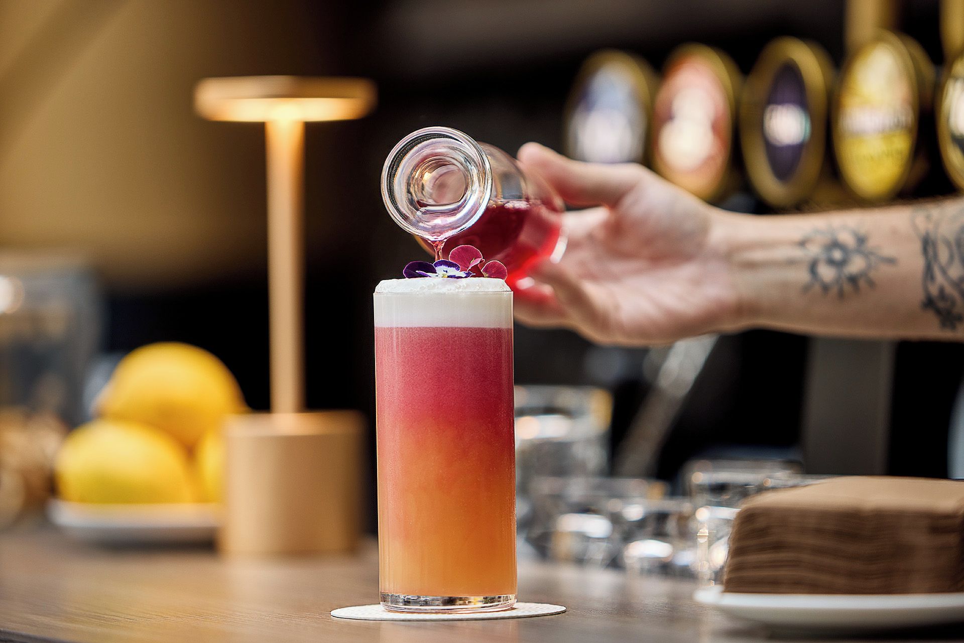 A tall glass with a gradient color, a hand pouring a drink into it, and a bar setting with bottles in the background.
