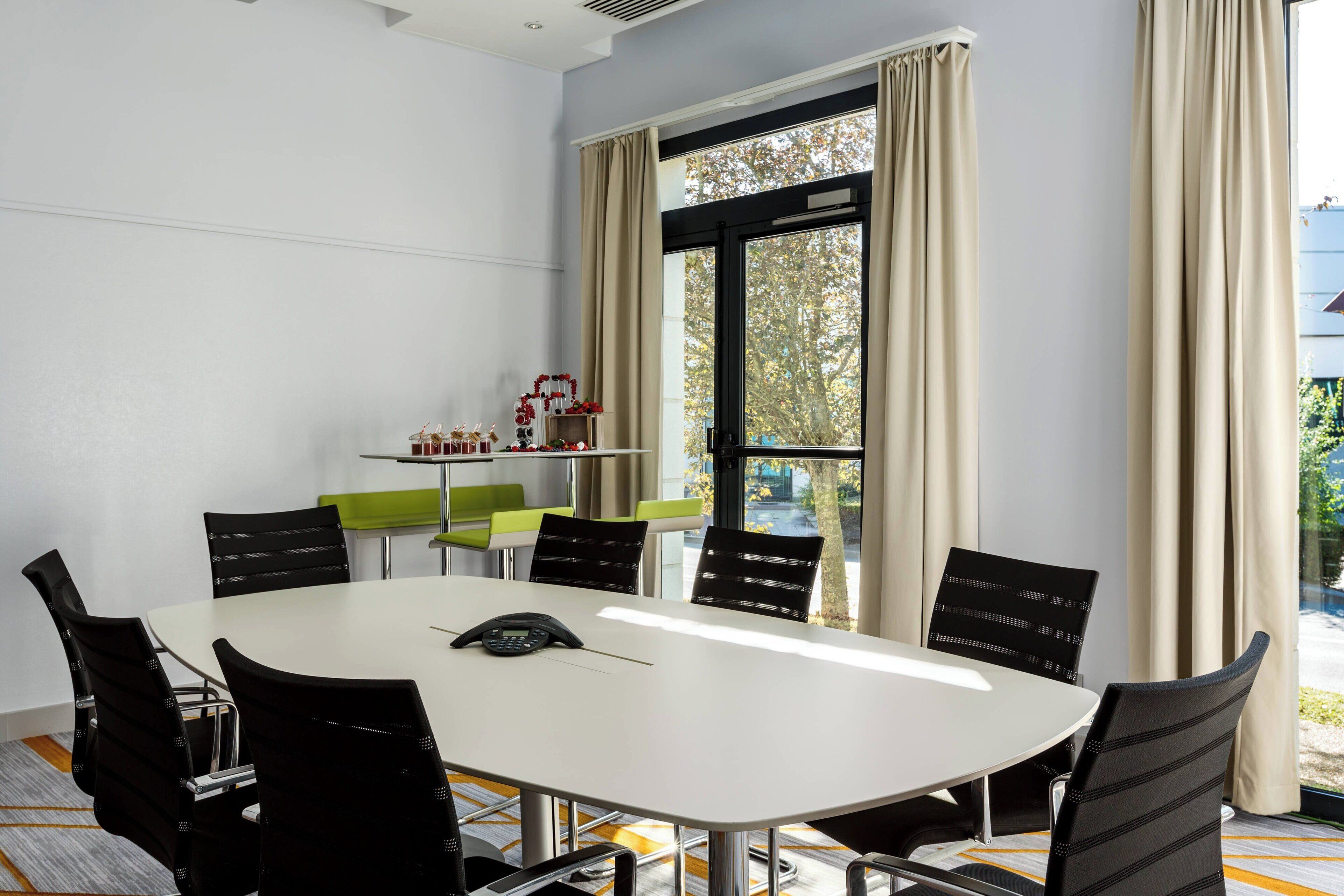 A conference room with a white table, black chairs, a phone on the table, and a window with curtains.