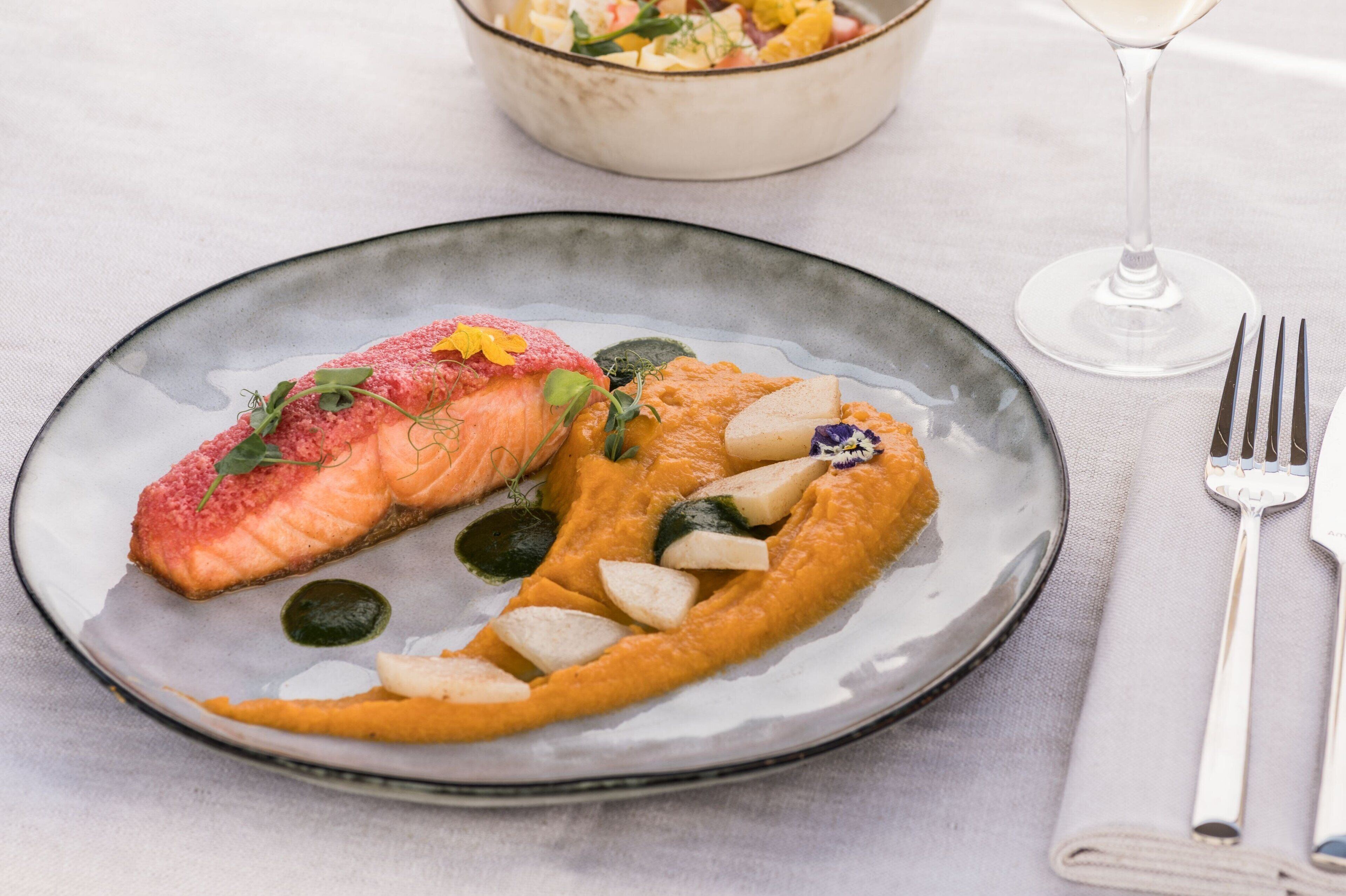 Un plat amb salmó i verdures sobre una taula, juntament amb una forquilla i un got d'aigua.