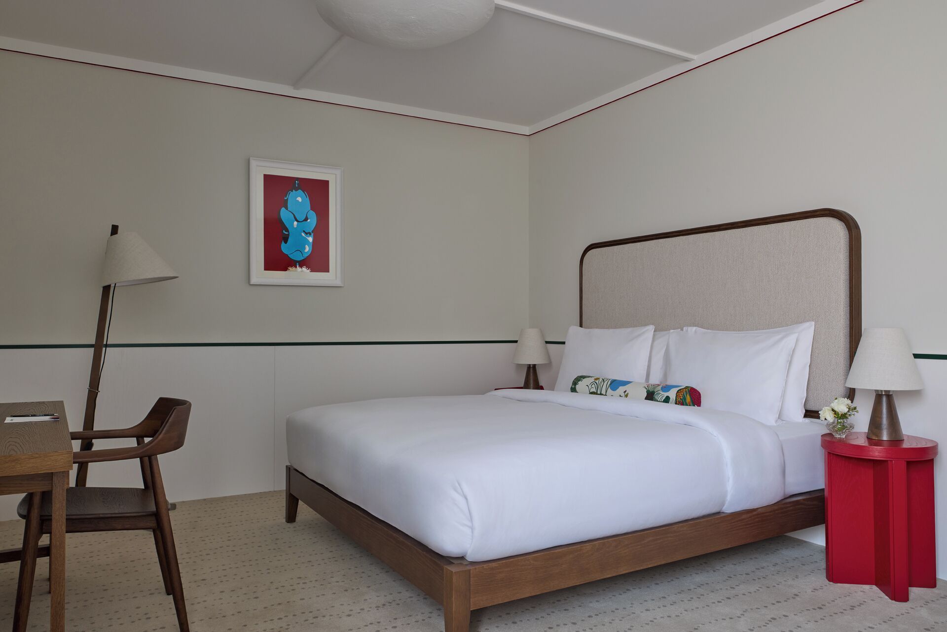 A hotel room with a bed, a desk, a chair, and a red side table.
