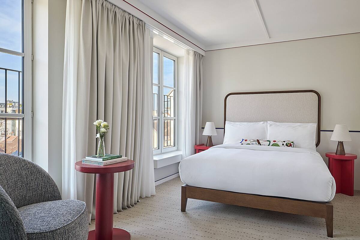 Habitación de hotel con cama, una silla, una mesita con un florero y ventana con cortinas.