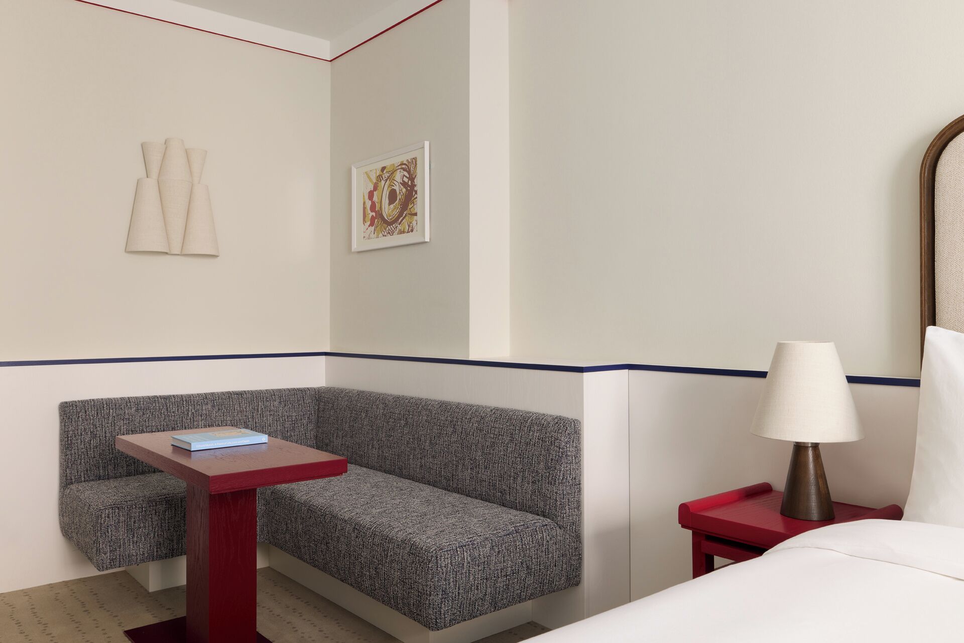 A hotel room with a gray upholstered bench, a small wooden table with a book, a red nightstand with a lamp, and a framed picture on the wall.