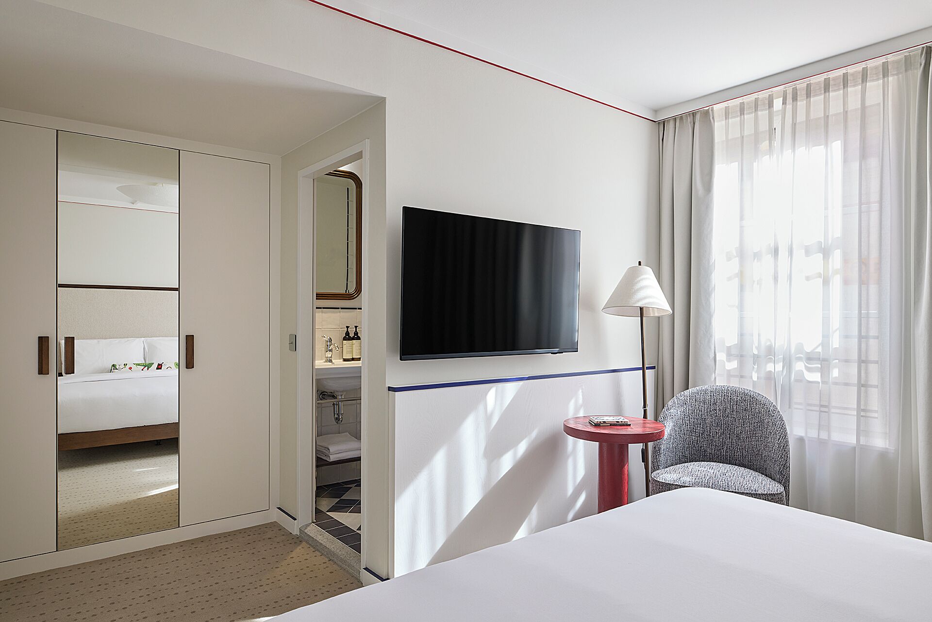 A hotel room with a bed, a TV mounted on the wall, a small table, a chair, a closet with a mirror, and a window with sheer curtains.