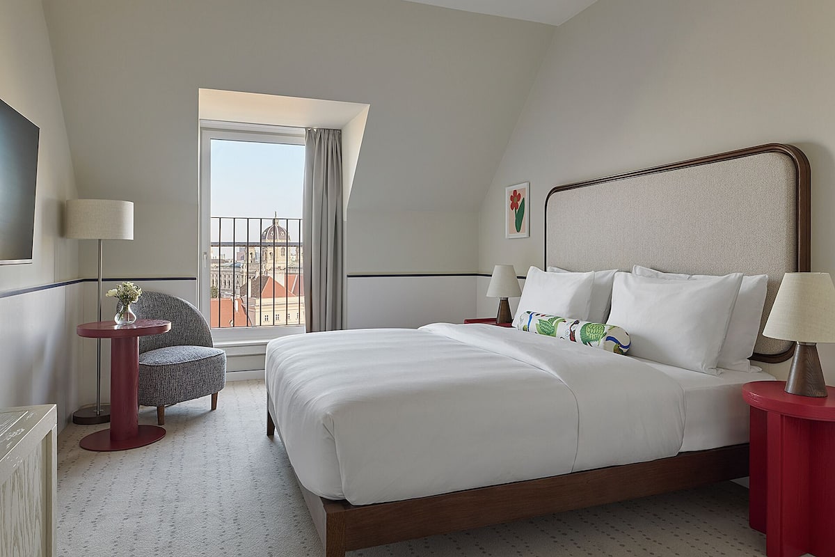 Habitación de hotel con cama, una silla, una mesa, una lámpara y ventana con vista a edificios.