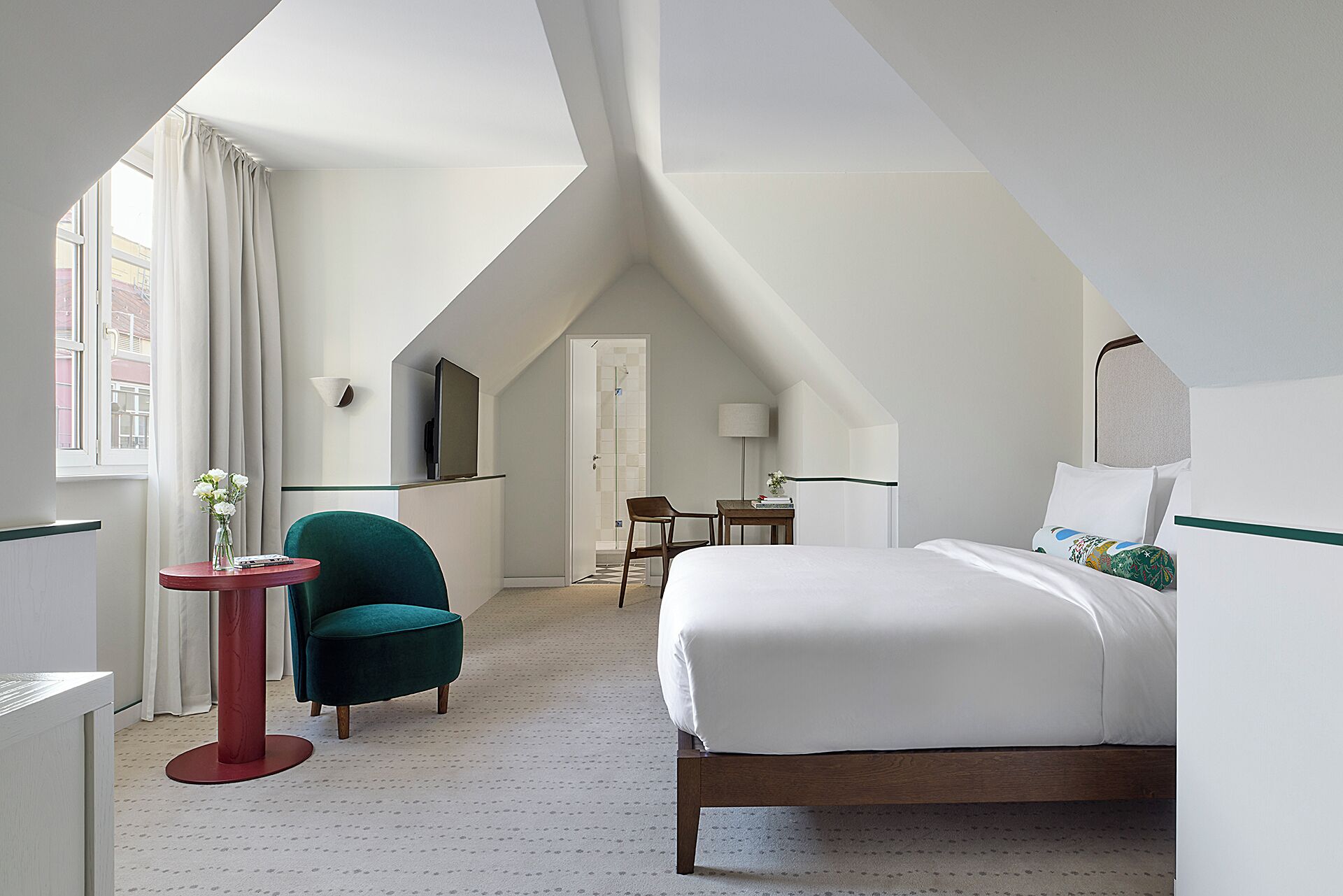Quarto de hotel com cama, poltrona, mesinha, televisão e janela com cortinas.