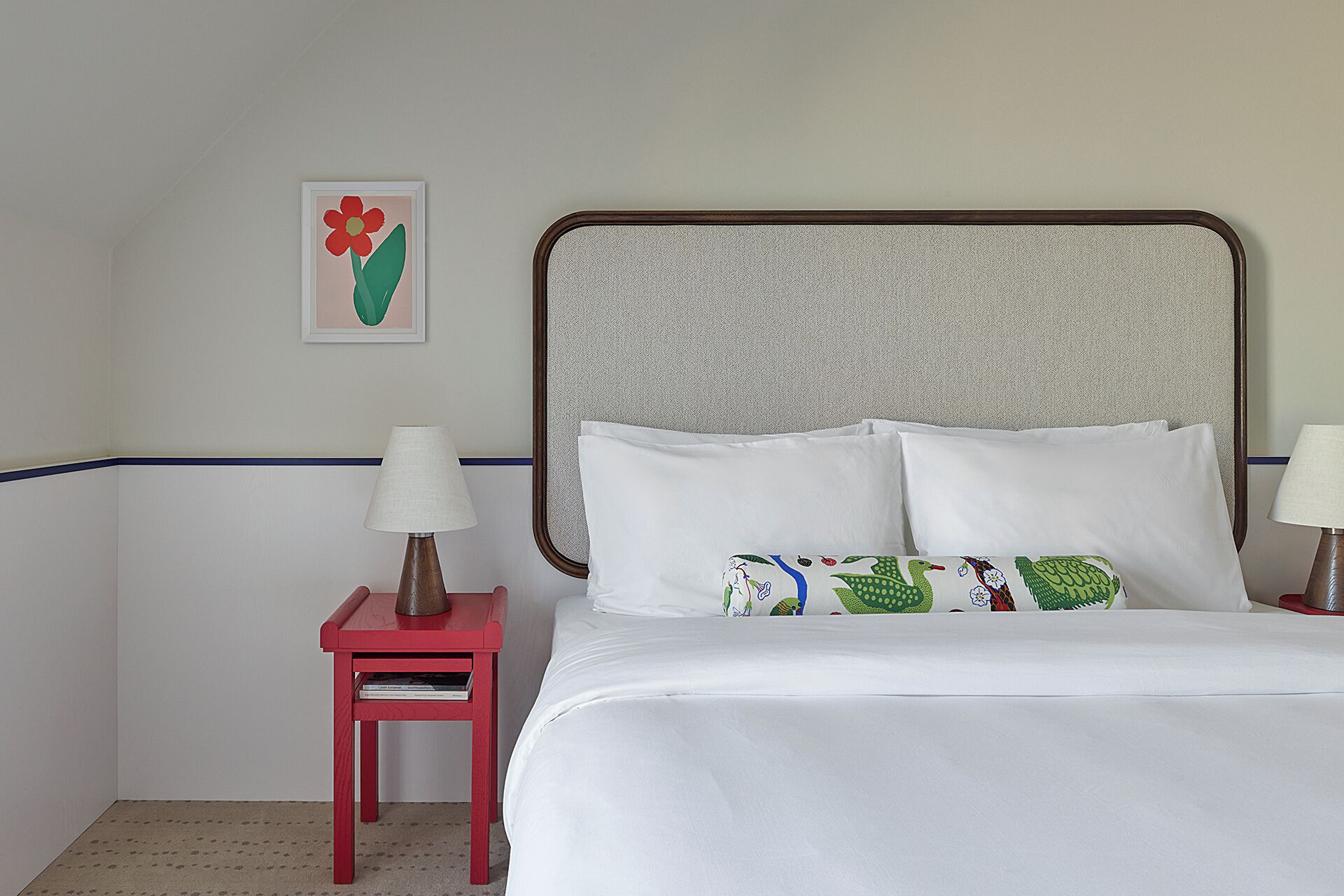 Una habitación de hotel con cama, almohadas, una mesita de noche con lámpara, un cuadro en la pared y una mesita auxiliar roja.