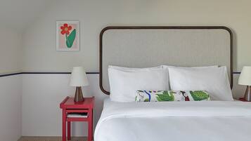 Una habitación de hotel con cama, almohadas, una mesita de noche con lámpara, un cuadro en la pared y una mesita auxiliar roja.