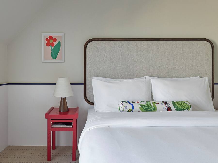 Una habitación de hotel con cama, almohadas, una mesita de noche con lámpara, un cuadro en la pared y una mesita auxiliar roja.