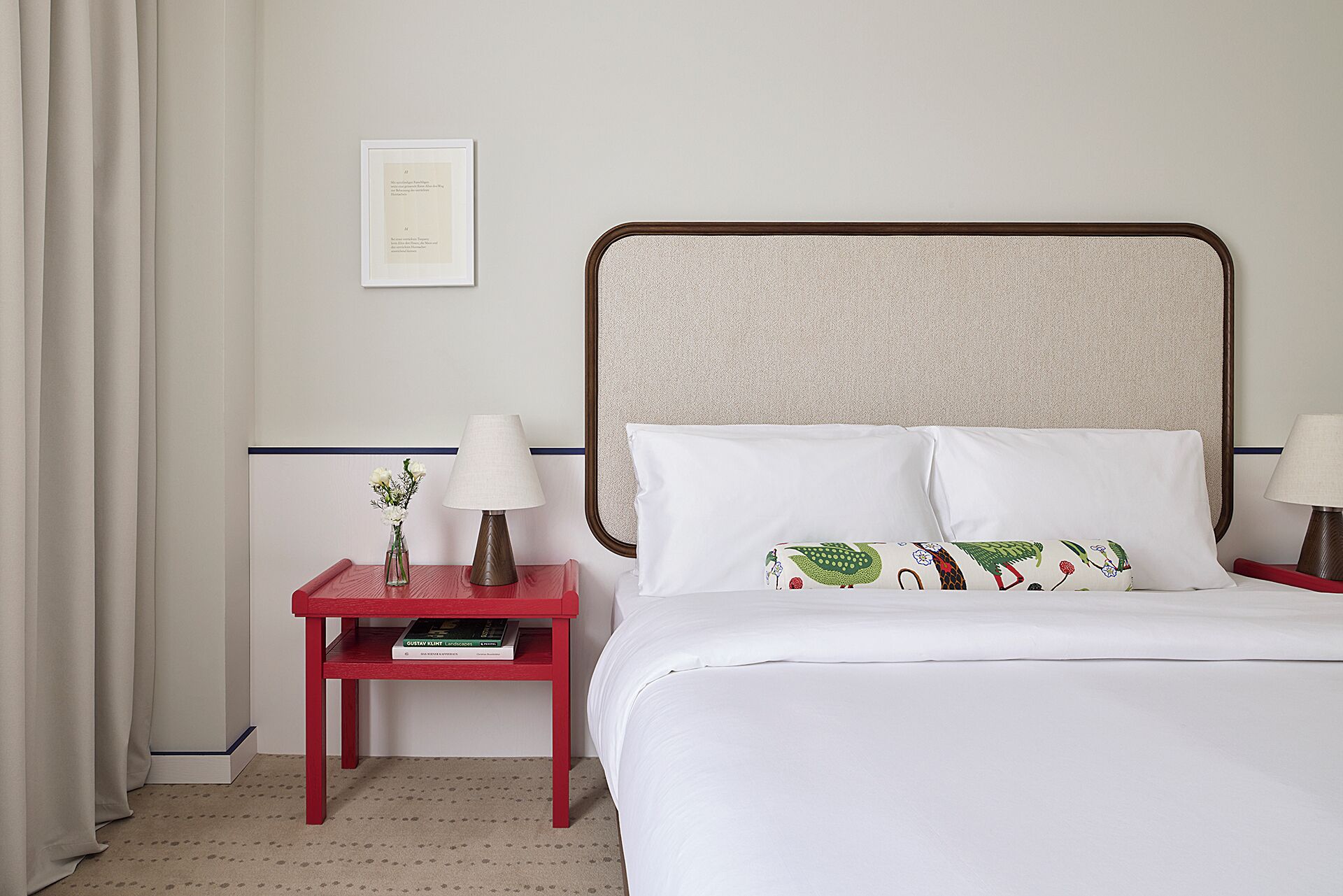 Une chambre d’hôtel comprenant un lit, deux tables de chevet, une petite table, un certificat encadré et un vase avec des fleurs.