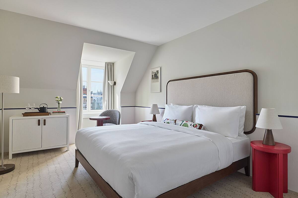 A hotel room with a bed, a desk, a chair, a window, and a lamp.