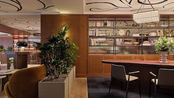 Un interno moderno con una scrivania di accoglienza in legno, una grande pianta in vaso, una zona pranzo con sedie e una libreria piena di libri e oggetti decorativi.
