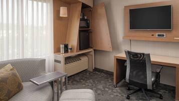 A compact hotel room with a sofa, a small desk, a chair, a TV mounted on the wall, and built-in appliances like a microwave and a mini-fridge.