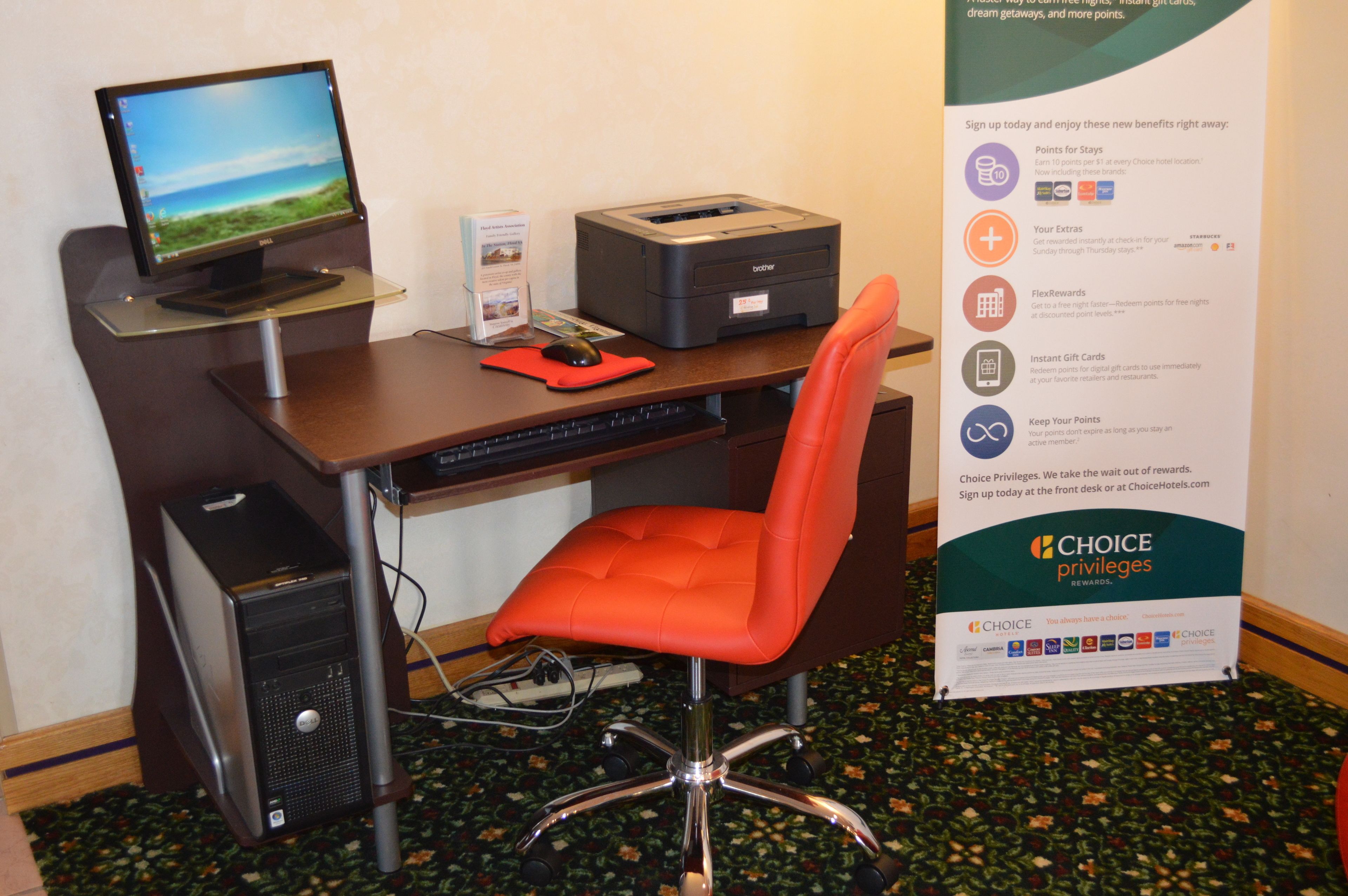 A hotel room with a desk, computer, printer, and a promotional sign for Choice Privileges.