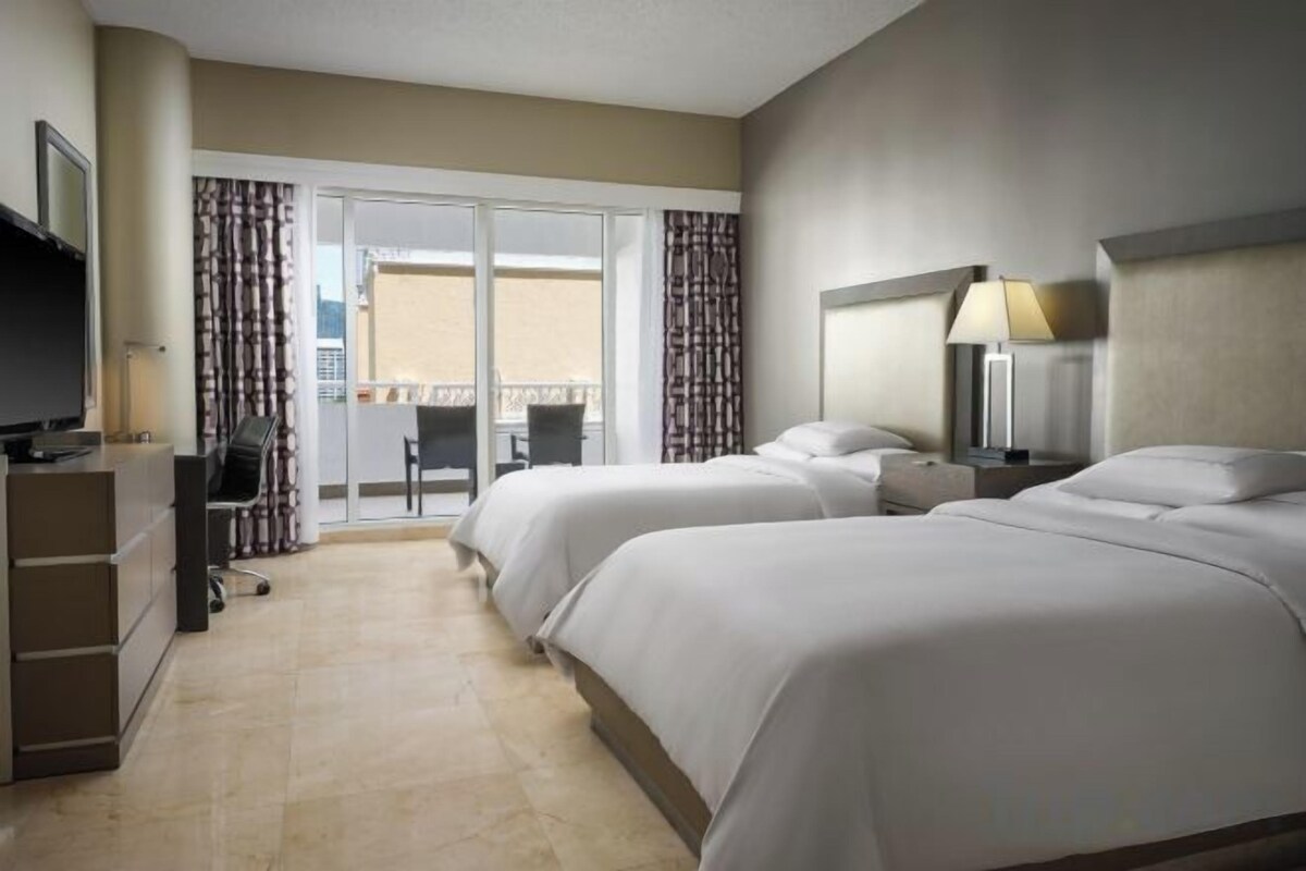 Quarto de hotel com duas camas, uma escrivaninha com cadeira, televisão e varanda com cortinas.