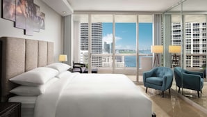 A modern hotel room with a large bed, a seating area with two armchairs, a nightstand, and a large window offering a city view. - DoubleTree by Hilton Grand Hotel Biscayne Bay (Miami)