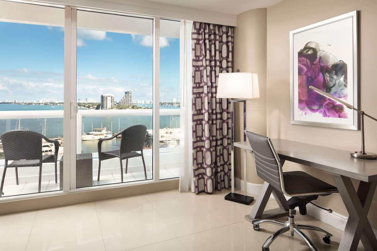 A hotel room with a large window overlooking a marina, a desk with a chair, and a framed artwork on the wall.