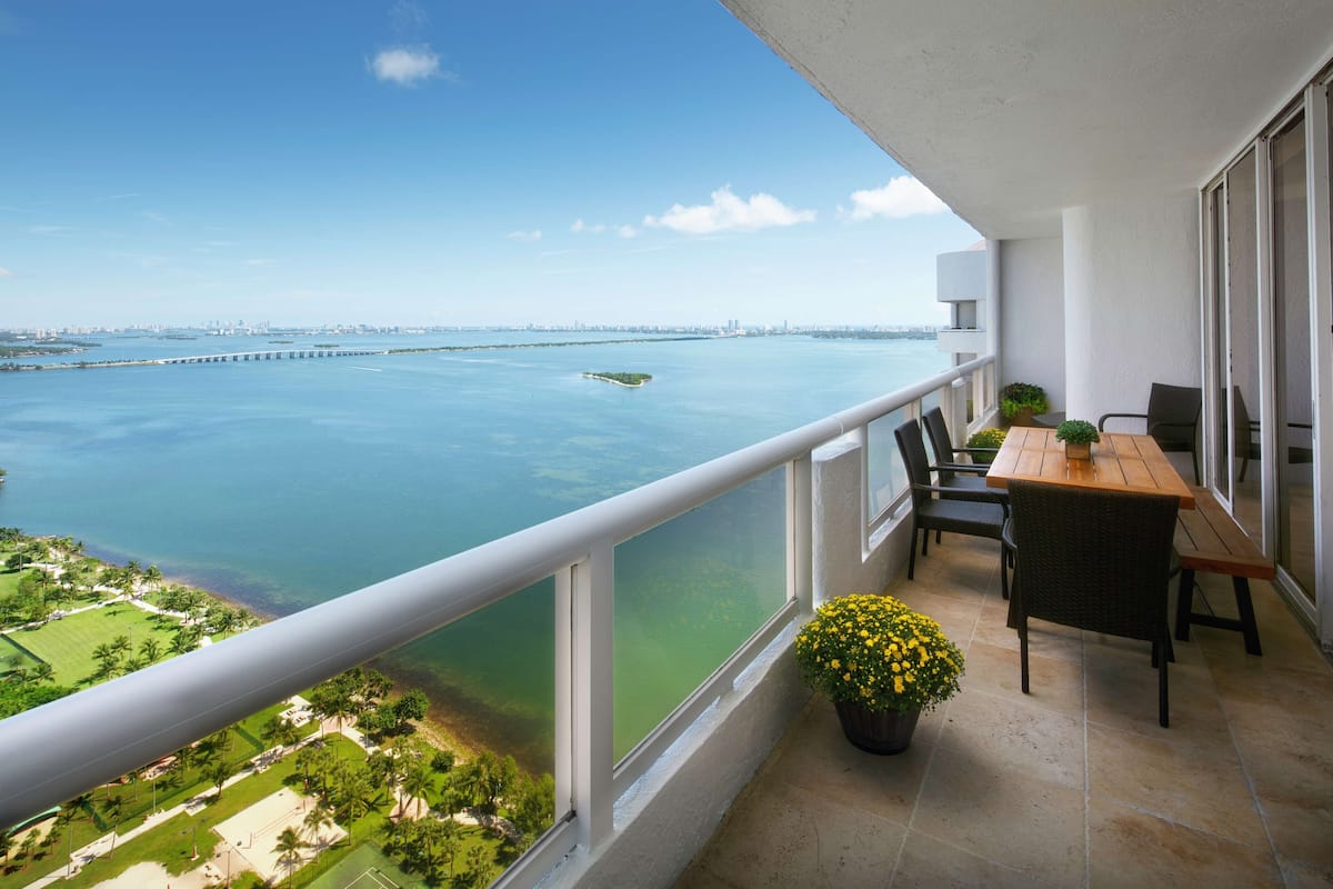 A balcony with a view of a large body of water, a wooden table, chairs, and a potted plant.