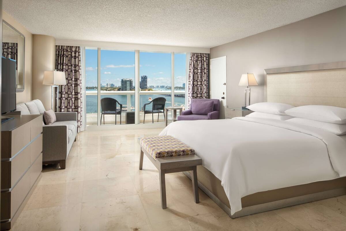 A modern hotel room with a large bed, a seating area with a chair and a table, a TV, and a view of the city and water.