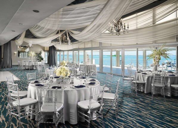 A banquet hall with round tables, clear chairs, and a view of the sea through large windows. - DoubleTree by Hilton Grand Hotel Biscayne Bay (Miami)