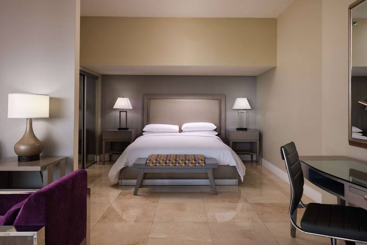 A hotel room with a bed, two bedside tables with lamps, a desk with a chair, and a large mirror.