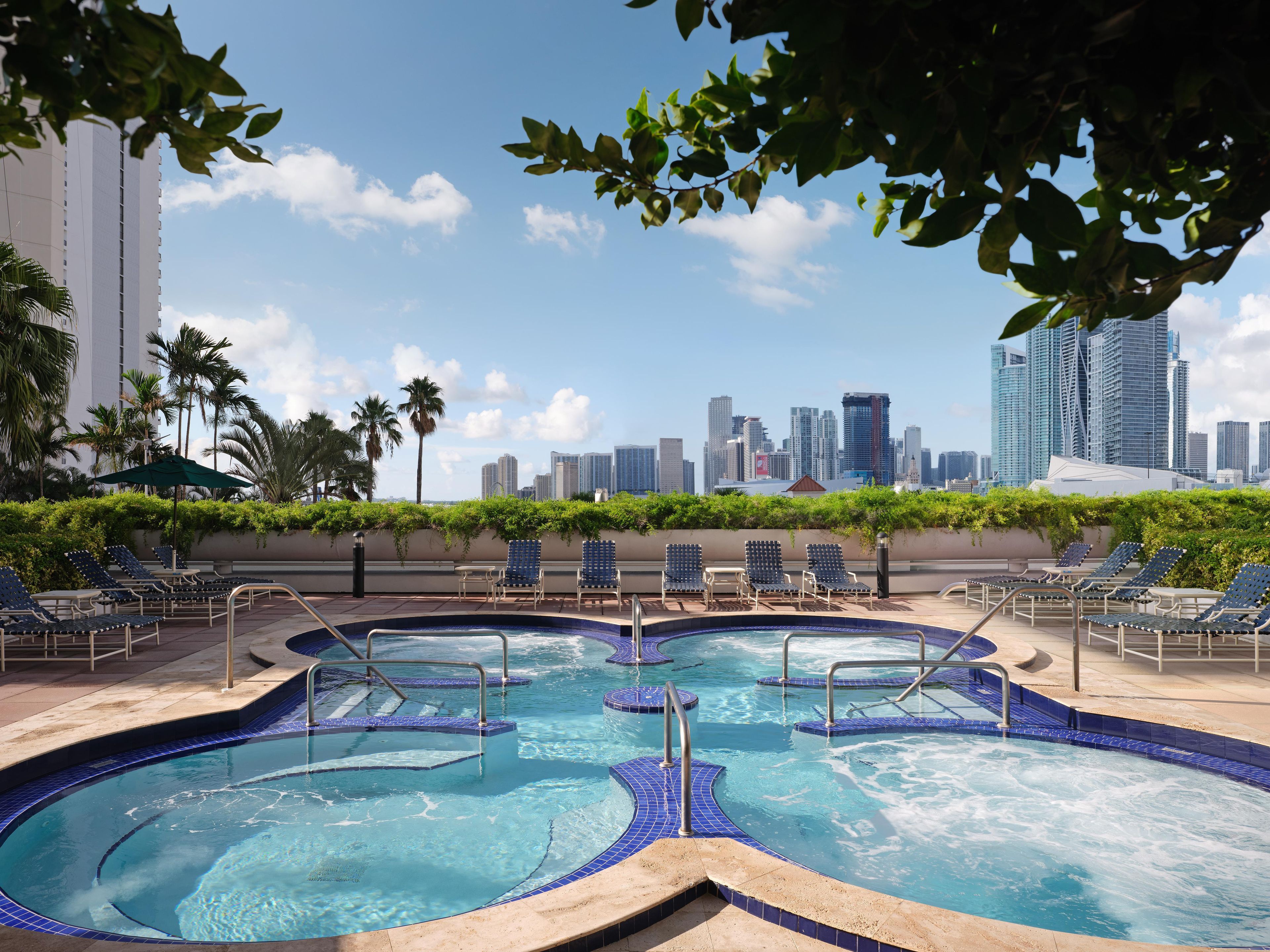 Una piscina en la azotea con varias pistas de nado, rodeada de sillones de descanso y palmeras, con un horizonte urbano al fondo.