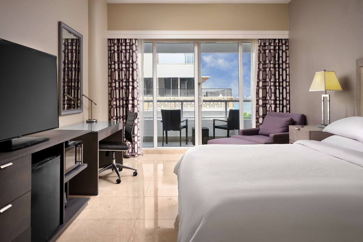 A hotel room with a bed, a desk with a chair, a TV, a lamp, and a view of the city through the balcony doors.