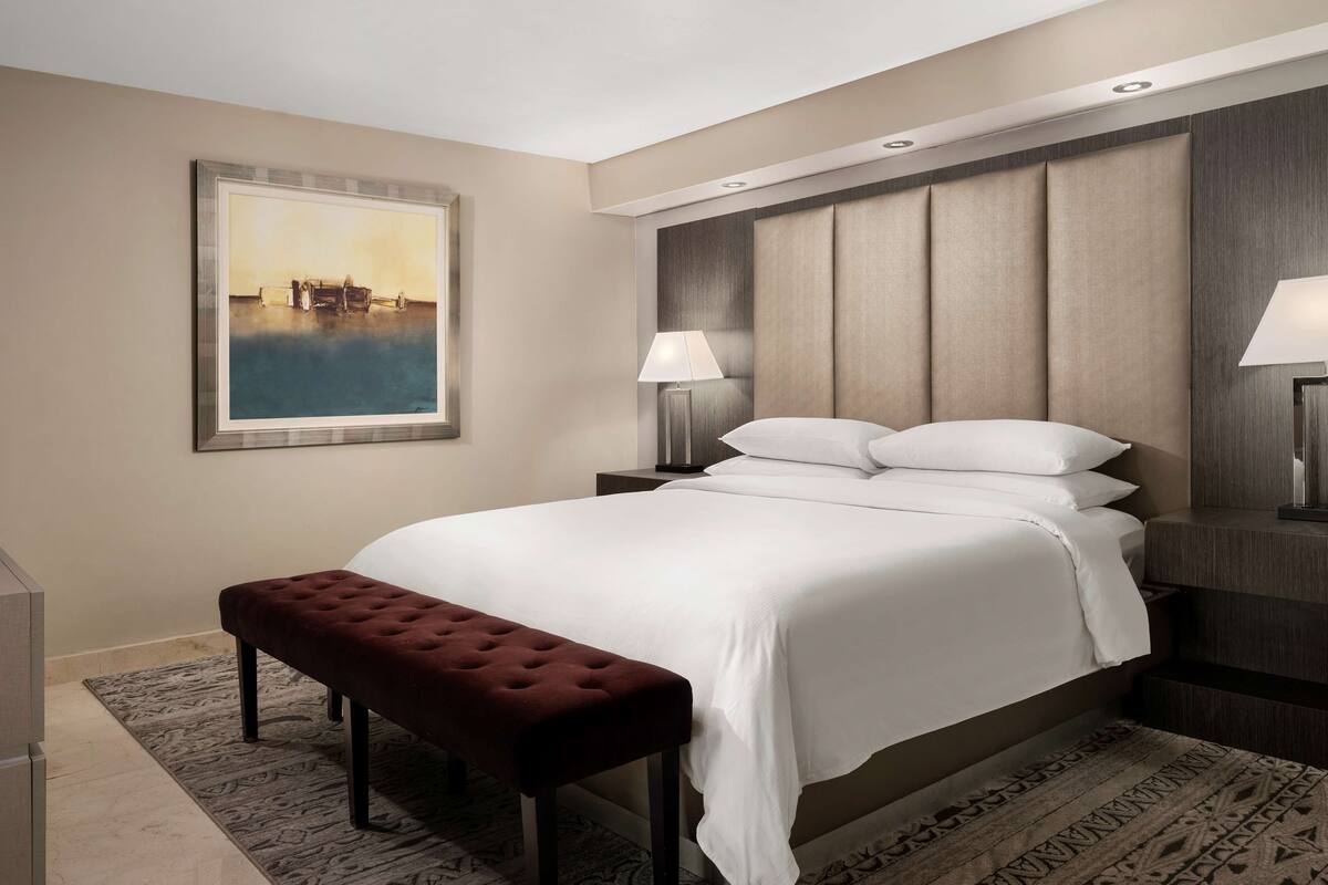 A hotel room with a large bed, a bench, a nightstand with a lamp, a painting on the wall, and a patterned rug.