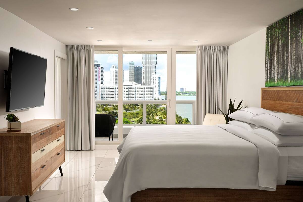 A modern hotel bedroom with a large bed, a TV, a wooden dresser, and a city view.