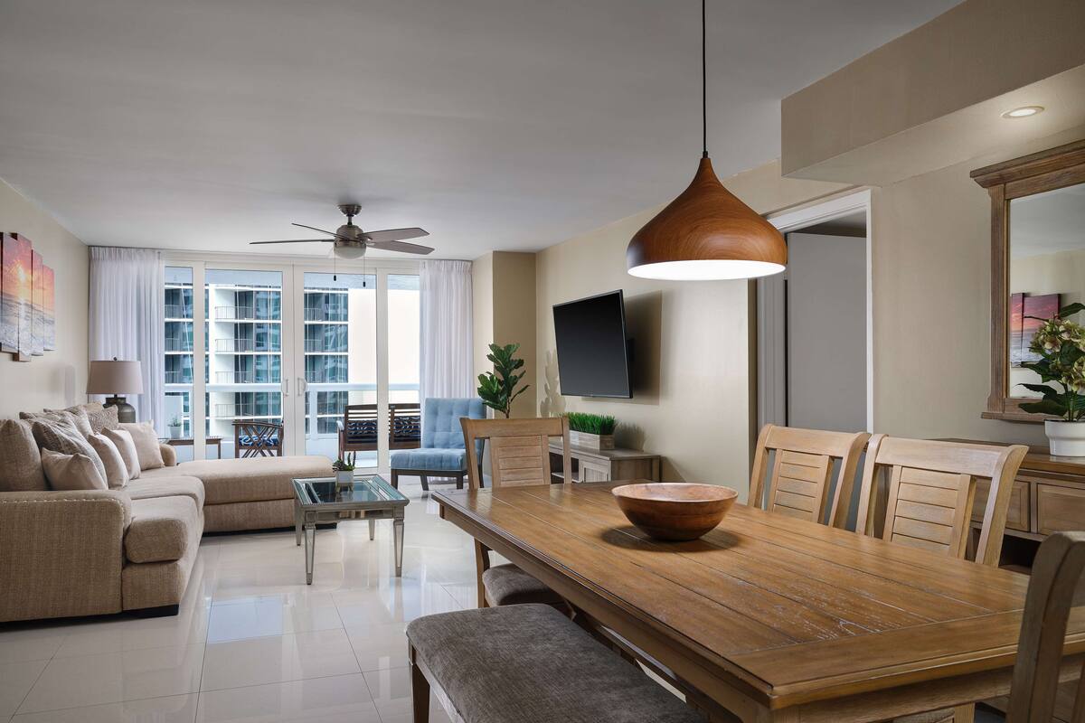A modern living room with a wooden dining table, beige sofas, and a ceiling fan.