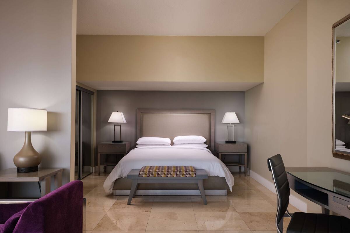 A hotel room with a large bed, two bedside tables with lamps, a desk with a chair, and a mirror on the wall.