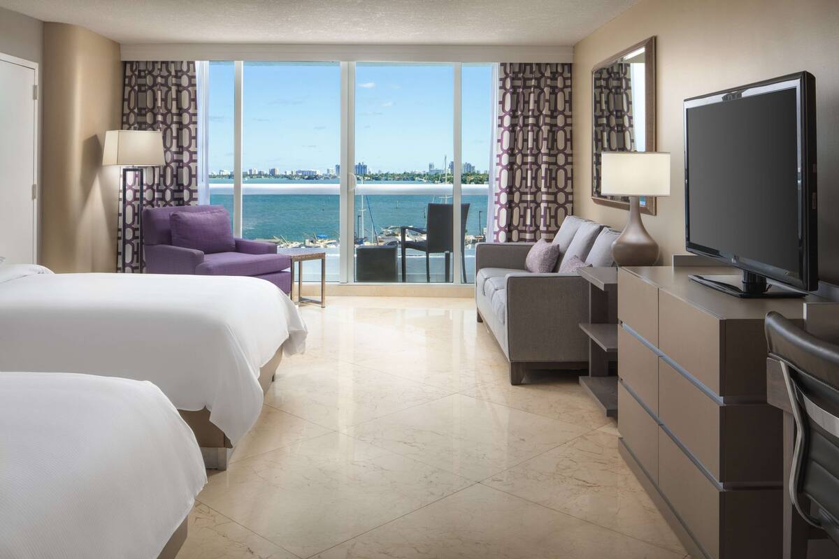 A hotel room with two beds, a sofa, a TV, and a view of the sea and city skyline.