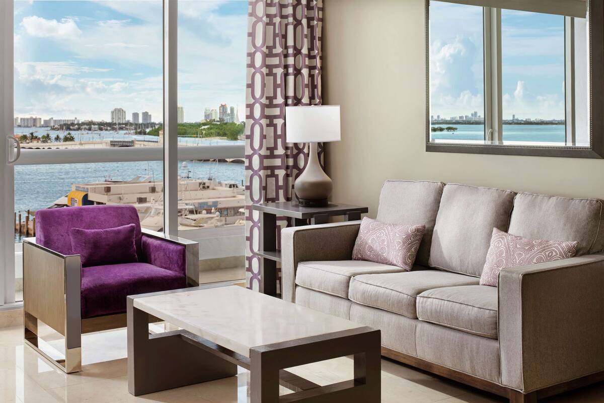 A modern living room with a view of a marina and city skyline, featuring a purple armchair, a beige sofa, and a minimalist coffee table.