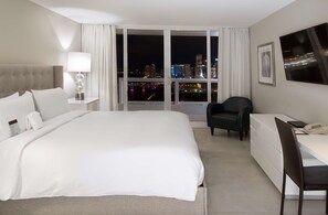 A hotel room with a large bed, a desk, a chair, and a view of a cityscape at night. - DoubleTree by Hilton Grand Hotel Biscayne Bay (Miami)