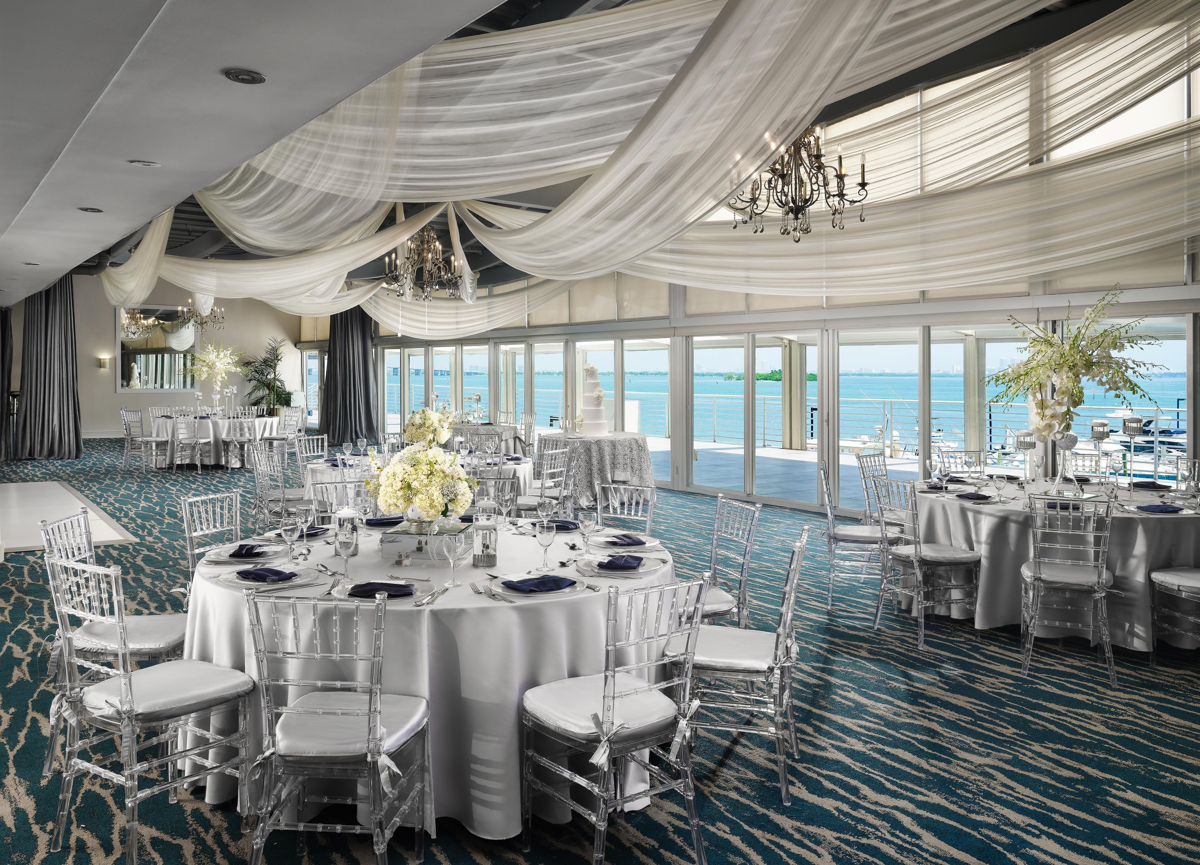 Un salón de banquetes con mesas redondas, sillas traslúcidas y vistas al mar.