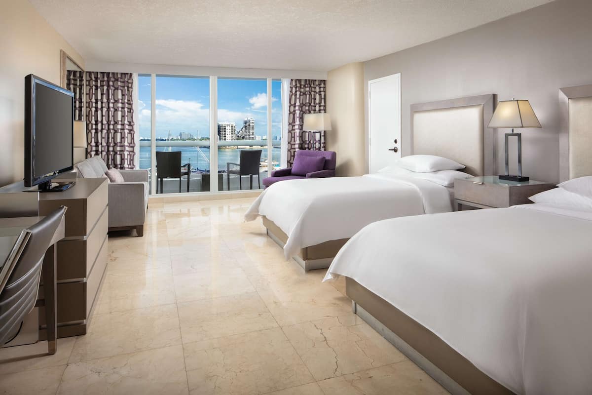 A hotel room with two beds, a TV, a desk, a chair, and a view of the city.
