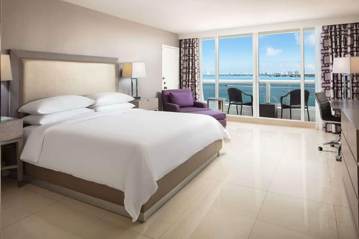 A modern hotel room with a large bed, a desk, a chair, and a view of the sea.