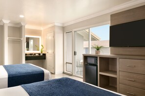 A hotel room with two beds, a kitchenette, a sliding glass door to a balcony, and a flat-screen TV mounted on the wall.