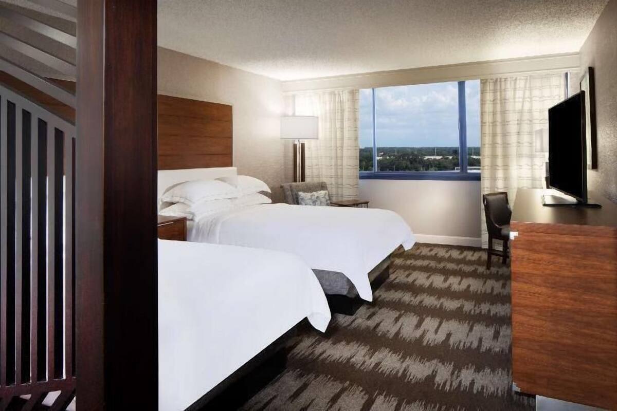A hotel room with two beds, a desk, a chair, a TV, and a large window with a view.