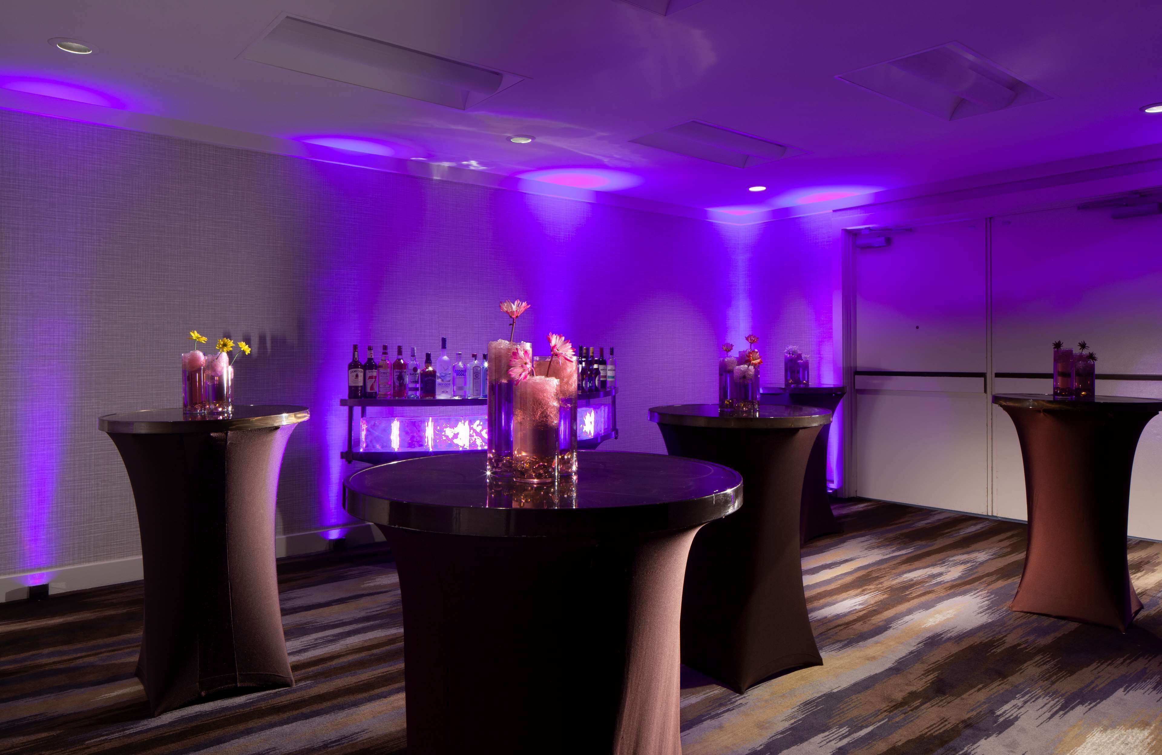 A room with three round tables, each with a vase of flowers. There is a bar area with bottles and glasses in the background.