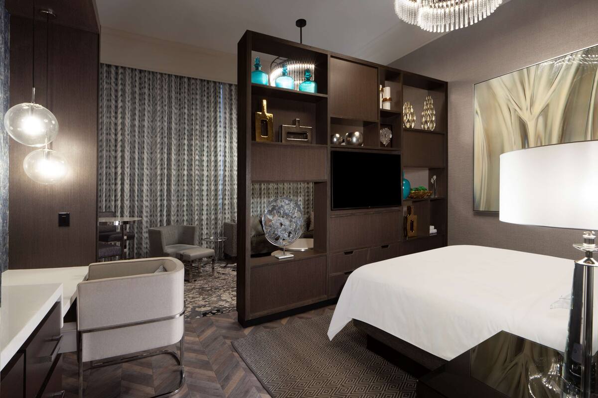 A modern hotel room with a large bed, a desk, a chair, a TV, and a minibar.