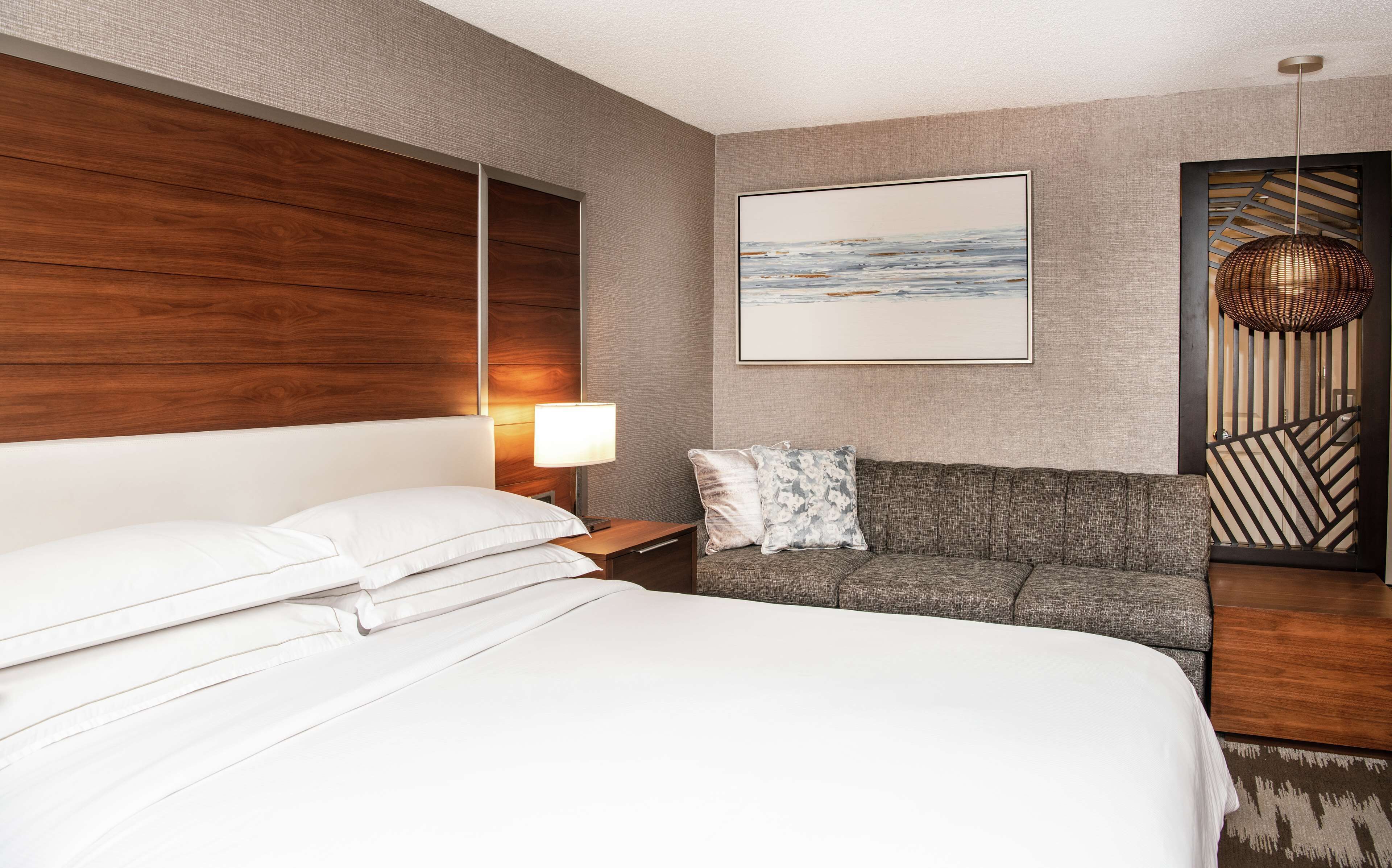 Una camera d'albergo con un letto grande, un divano grigio, una testiera in legno, un'opera d'arte incorniciata appesa al muro e un lampadario.
