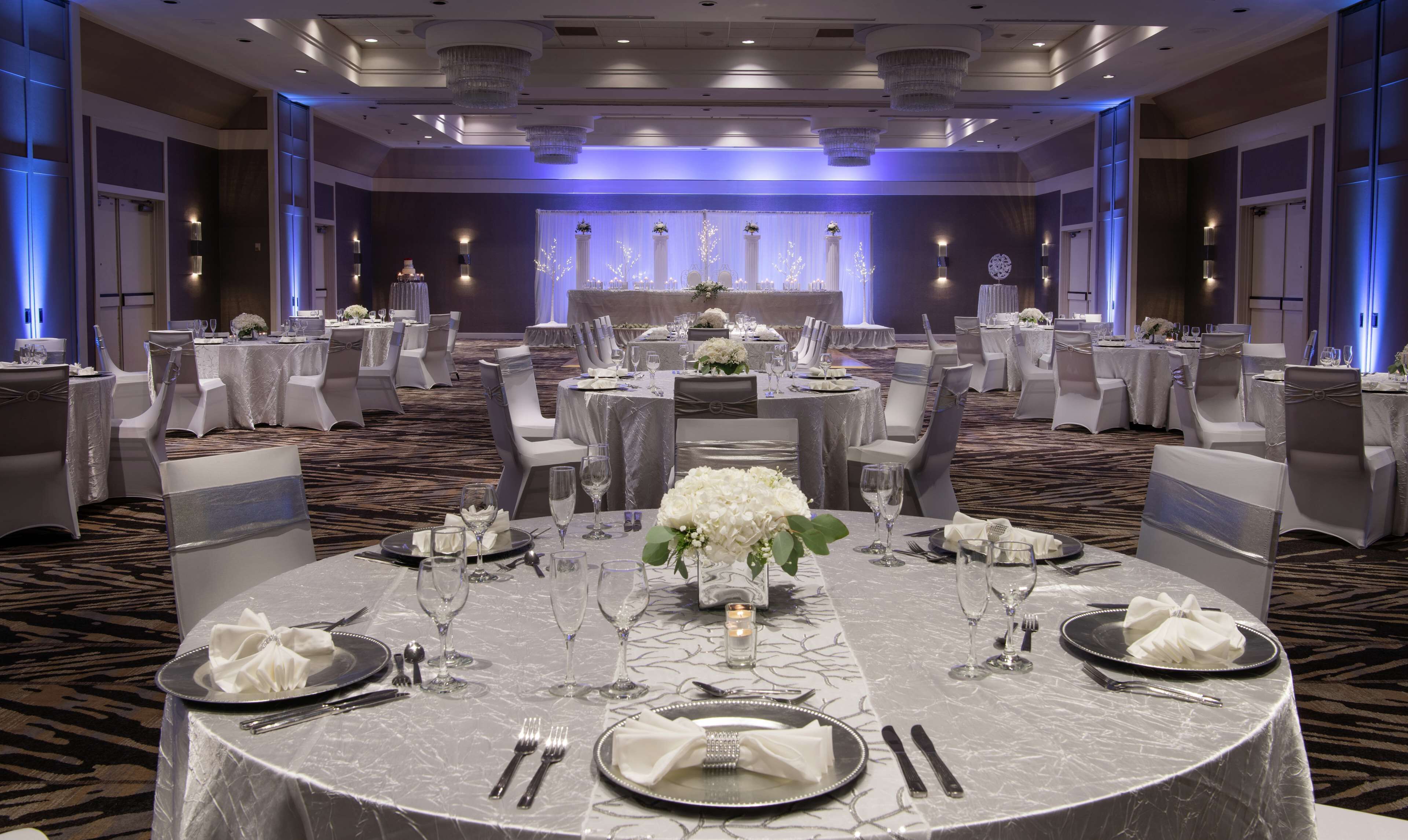 Un salón de banquetes con mesas redondas dispuestas para un evento formal, con manteles blancos, cubertería y centros de mesa florales.