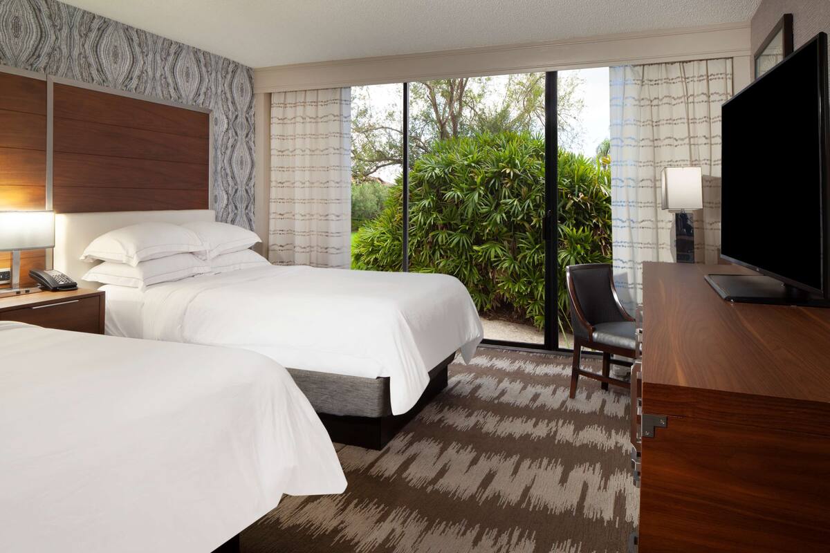 A hotel room with two beds, a desk, a TV, and a view of greenery outside.