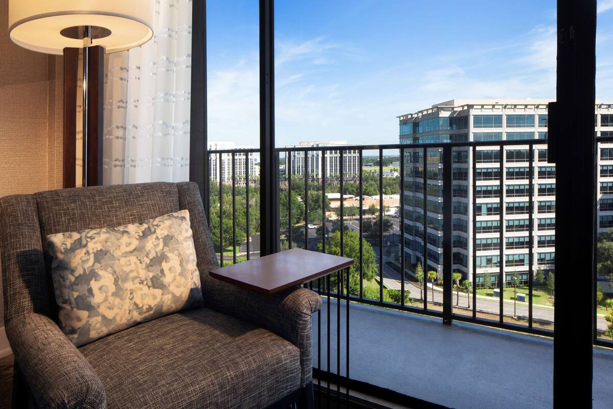 A modern hotel room with a large window, a sofa with a patterned cushion, and a small table.