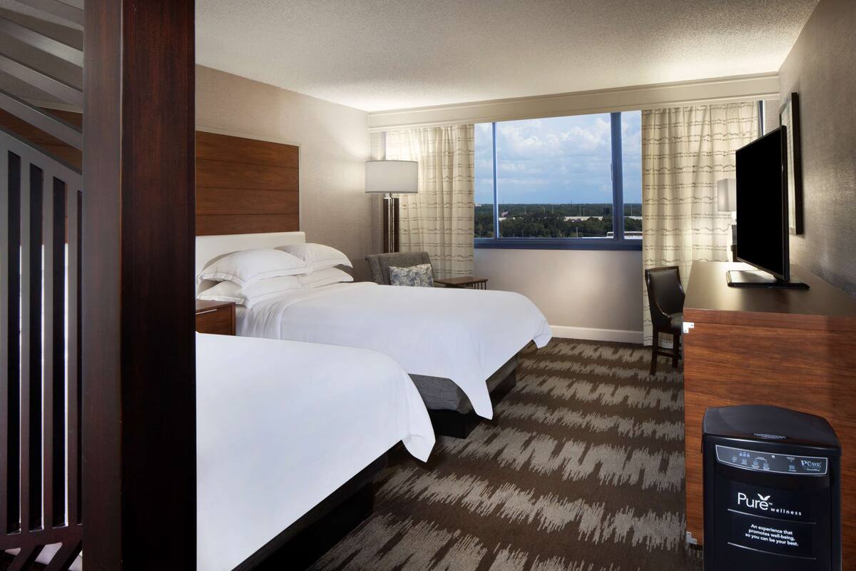 A hotel room with two beds, a desk, a chair, a TV, and a view of the city.